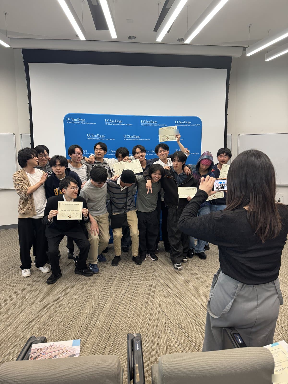修了式で、UCサンディエゴのパネルの前で修了証書を手に記念撮影をする参加学生たち