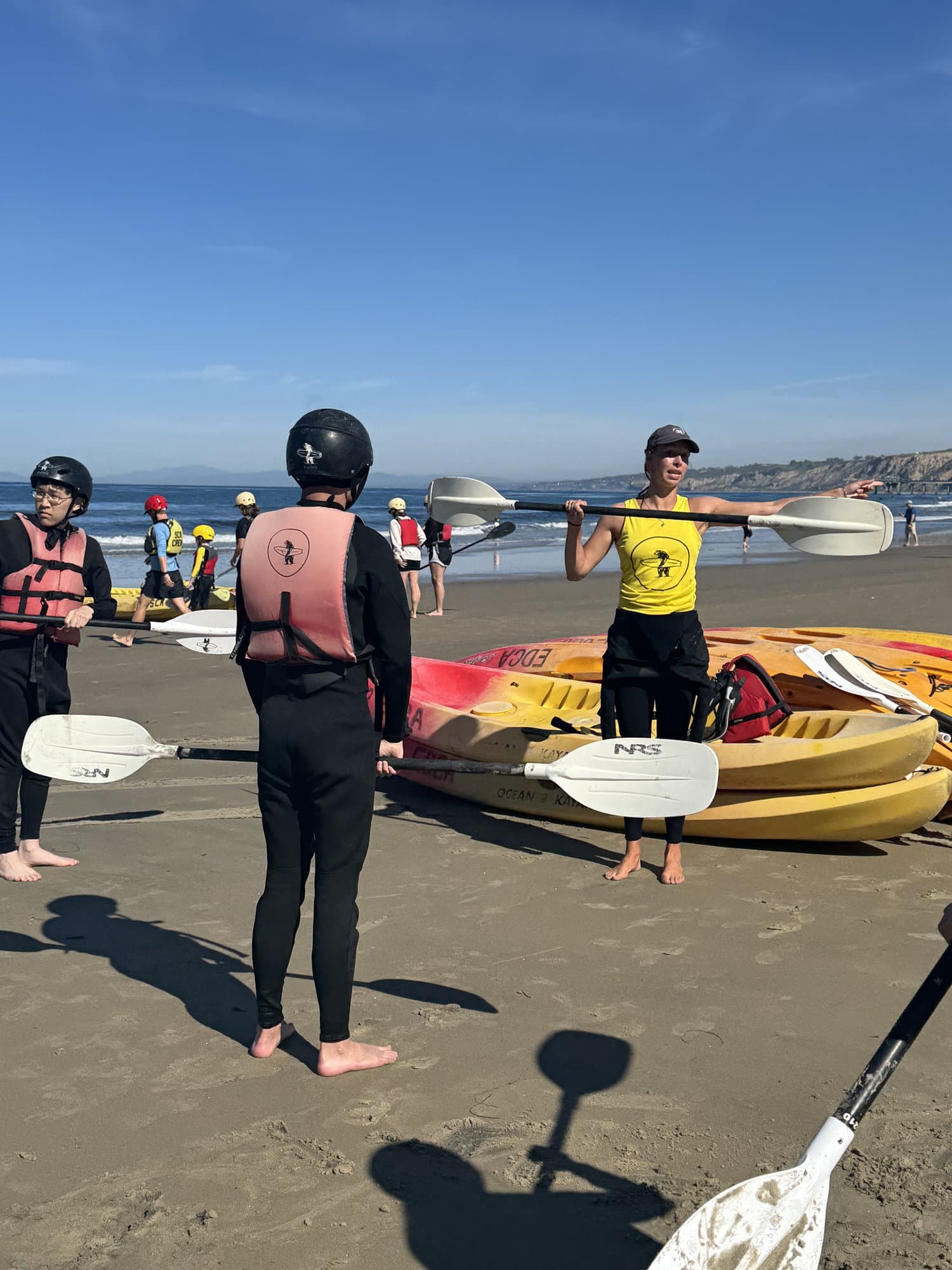 過去の参加者のカヤックレッスン。浜辺で、ヘルメットとライフジャケットを着けた学生にパドルの使い方を教えるインストラクター